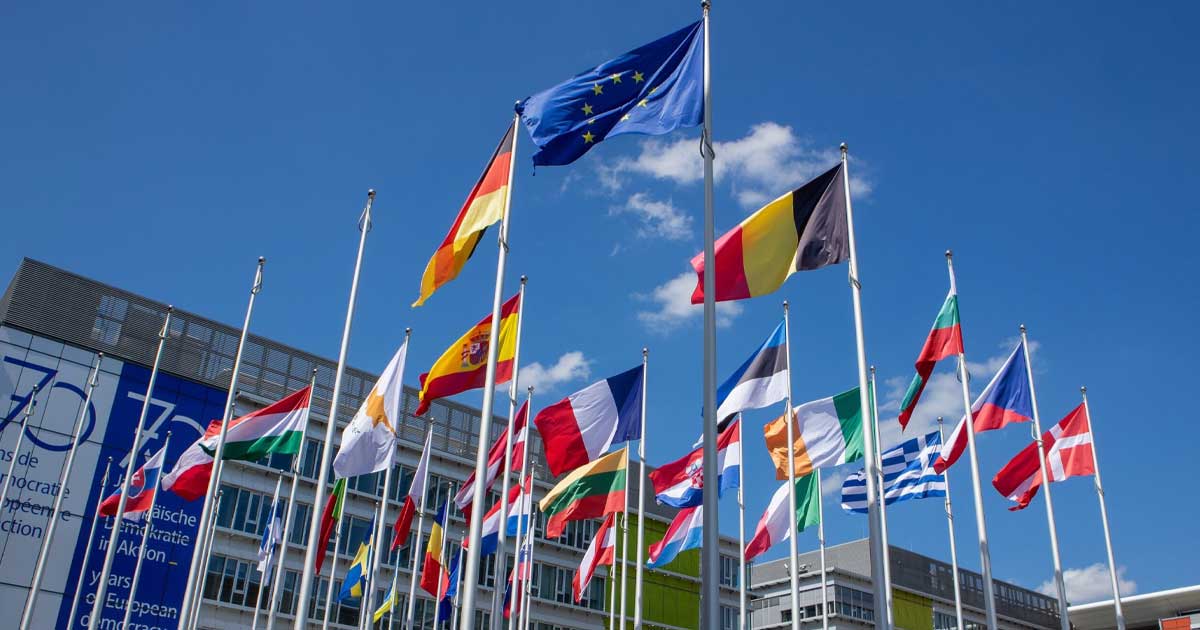 Clădirea Comisiei Europene din Bruxelles cu steaguri ale statelor membre în fața intrării, sub un cer senin